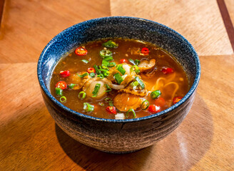 Spicy Asian Scallop Noodle Soup in a Blue Bowl topped with chili peppers, and green scallions on a Wooden Table side view of Asian food
