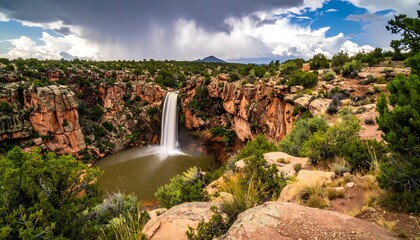 A dramatic waterfall cascades into a serene pool surrounded by rugged, rocky cliffs and lush greenery under a stormy sky