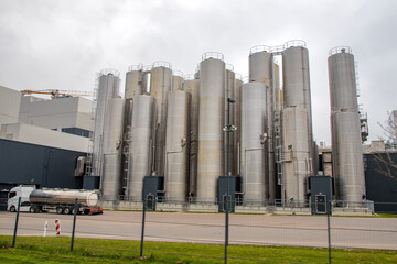 A row of tall, metallic storage silos at an industrial facility, with a tanker truck parked in front, under a cloudy sky.