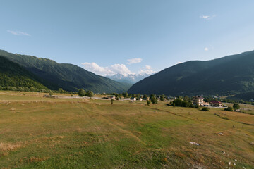 A tranquil countryside landscape unfolds across a wide valley framed by gentle hills. Sparse trees dot the fields beneath a clear blue sky, evoking peaceful, timeless natural beauty.