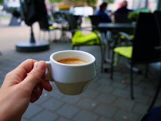 Hand holding white coffee cup in outdoor café – green cushions, tables, city street, urban vibe, social moment, casual lifestyle, public space, relaxed atmosphere, people and architecture