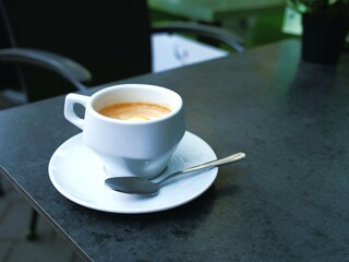 Hand holding white coffee cup in outdoor café – green cushions, tables, city street, urban vibe, social moment, casual lifestyle, public space, relaxed atmosphere, people and architecture