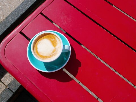 Cappuccino or latte with foam art on red wooden table &ndash; white cup, turquoise saucer, outdoor caf&eacute; scene, natural light, cozy coffee moment, vibrant color contrast, relaxing atmosphere.