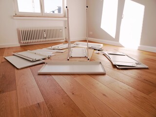 Flat-pack furniture assembly in bright room – white panels, metal rods, screws, mirrored wardrobe, wooden floor, DIY setup, home interior, cabinet construction, hardware layout.