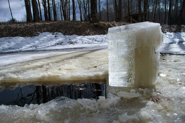 A large, clear ice block stands tall in a partially frozen river during the late winter or early...