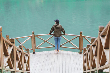 Woman in a jacket and cap standing on a wooden platform by a turquoise lake, enjoying peaceful nature and solitude, concept of travel and mindfulness