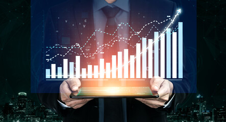 A businessman stands in a modern office, holding a tablet displaying financial graphs and data...