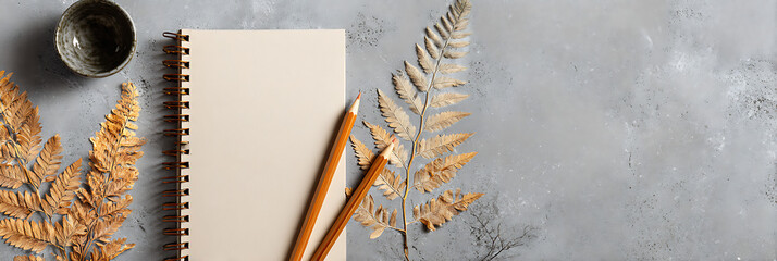 Flat lay of a spiral-bound notebook and two pencils next to dried brown ferns, resting on a textured gray concrete background, symbolizing writing, nature, and creative planning.
