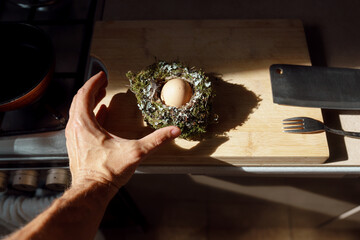 A hand reaching for an egg from a bird's nest. A bird's nest in the table at the background. Kitchen interior.