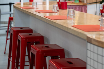 A retro-style diner interior with stackable red metal stools at a white counter topped in cream. Cloth placemats and clear drinking glasses line the counter, evoking a classic, nostalgic charm.
