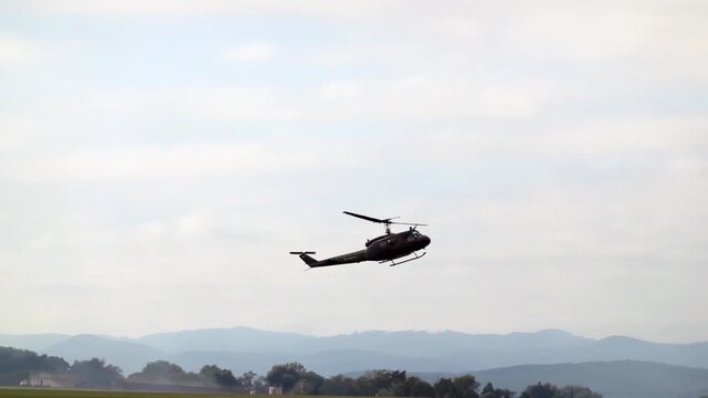 A retro UH-1 helicopter hovers above the ground and a smoking aerobatic plane approaches it from the right, slow motion