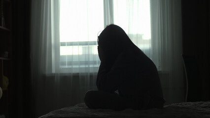 Silhouette of a millennial woman sitting on a bed in the dark bedroom, depression, pain. Portrait of Unhappy sad woman sitting against window and thinking about her life