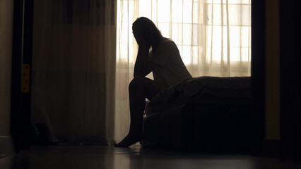 Silhouette of unrecognized woman sitting on a bed in the dark bedroom, depression, pain. Portrait of Unhappy sad woman sitting against window and thinking about her life