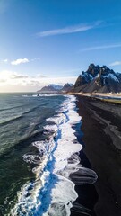 Aerial view of a dramatic black sand beach meeting a turquoise ocean under a vibrant blue sky, snow-capped mountains in the background