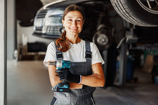 A smiling female mechanic confidently holds a power tool within a bustling auto repair shop environment - Powered by Adobe