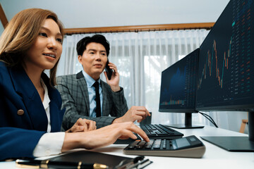 Naklejka premium Smiling Asian businessman calling to broker dynamic stock exchange database with woman business partner pointing to screens comparing profit value online application program at home office. Infobahn.