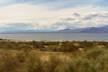 Obraz premium A motion-blurred desert landscape featuring a vast lake and distant mountains under a partly cloudy sky.