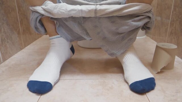 Man sits on toilet sink in restroom and impatiently twitches his legs. Feet of man sitting on toilet in white socks and with his underwear down. Maybe man can not poop and suffer from constipation