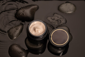 sleek jar of creamy skincare product sits on dark stones surrounded by water droplets. The setting reflects a serene atmosphere highlighting the quality and elegance of the cosmetic.