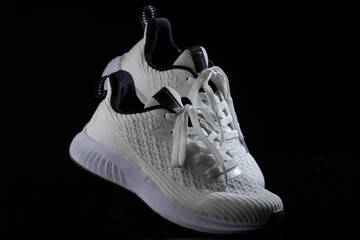 pair of white sneakers featuring a textured upper and black accents rests on a dark surface. The laces are untied adding a casual look. The sneakers highlight contemporary athletic footwear design.