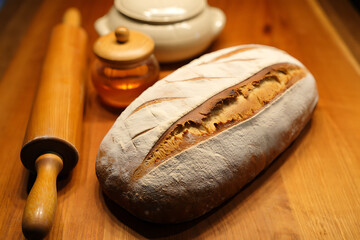 A rustic loaf of freshly baked bread, dusted with flour, sits on a wooden surface next to a rolling pin and a small jar of honey