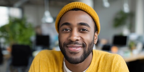 Smiling man in yellow sweater and beanie enjoying time in a modern office setting with plants and computers around.