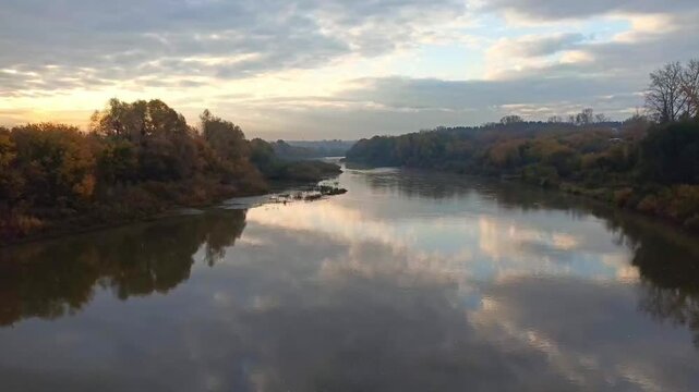 Reflections of clouds and trees along the banks of a wide river on an early autumn morning. Time-lapse. Inya River, Novosibirsk, Siberia, Russia.