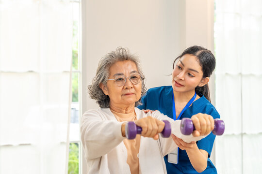 Asian woman physiotherapist caregiver teaching senior female patient exercise weight lifting dumbbell in bedroom. Elderly medical healthcare and wellness services, physical therapy and nursing home.