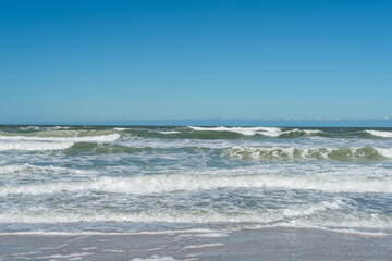 A view of a sandy beach on a clear sunny day. The sky is blue and the waves are rolling on shore with multiple waves. The tropical seashore has crashing white foam waves. The splashing surf has motion