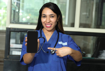 Indian happy female nurse showing mobile phone scree, while standing in hospital. Beautiful young nurse woman wearing uniform and stethoscope. Concept Healthcare