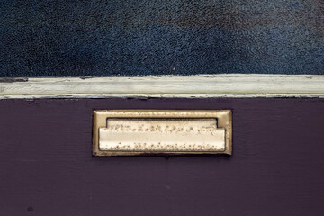 A vintage exterior wooden purple door with beige trim. There's a small brass post letter, or mail slot, in the center of the old door. The brass is rusty.