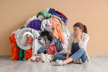 Troubled young woman near washing machine with pile of dirty laundry on beige background