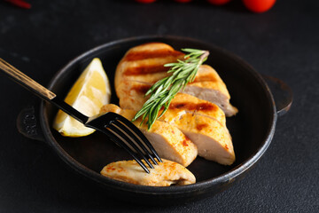Frying pan with cut grilled chicken breast, rosemary and lemon slice on black grunge background