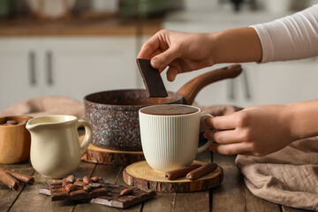Woman adding chocolate bar to cup of melted chocolate on table in kitchen. Closeup