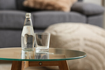 Bottle of fresh water and glass on table against stylish sofa in room