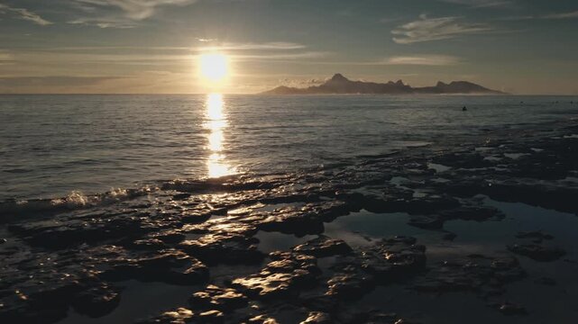Golden sunset reflecting on the wet volcanic rocks of the Tahiti coastline at dusk, creating a tranquil and scenic view with Moorea island in the background