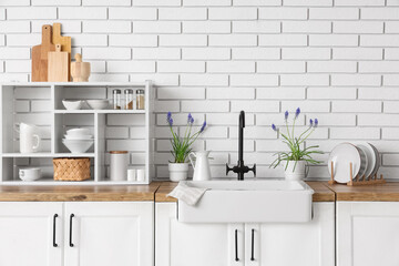 Stylish sink, beautiful Muscari flowers and shelving unit with kitchen utensils on counter near light brick wall