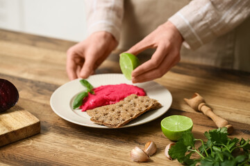 Woman squeezing lime onto tasty beet hummus at table in kitchen