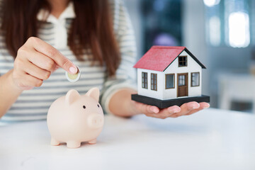 saving for home, mortgage down payment, real estate investment, financial planning, and personal money management, Woman saving coin in a piggy bank with house model beside © Sumala