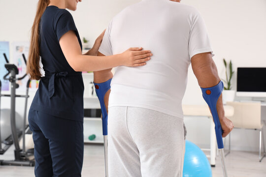 Female therapist and senior man with crutches walking in rehabilitation center, back view