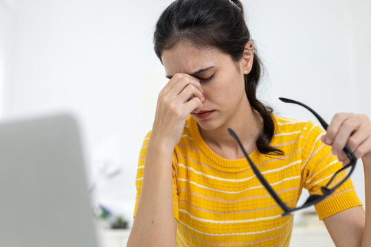 Tired Asian women removing glasses and rubbing eyes while working, expressing stress, digital eye strain, burnout, or fatigue from screen time and overwork.