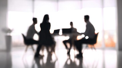 A blurred image of four individuals seated around a table in a modern office setting, engaged in a discussion or meeting.