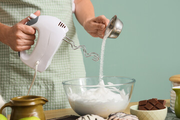 Woman whipping egg whites with sugar and mixer at table on green background, closeup