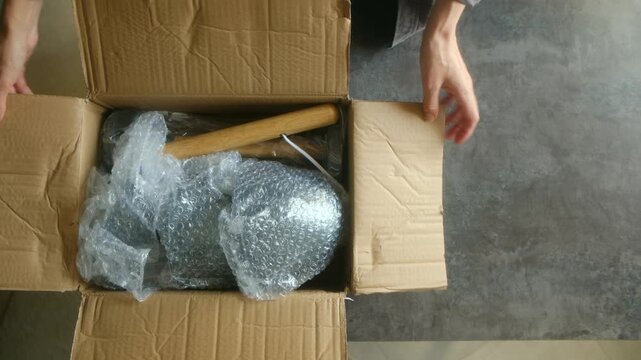 Top view woman hands unboxing new modern chandelier from cardboard box, removing bubble wrap from individual parts before starting assembly process at home. Woman unboxing assembling modern chandelier