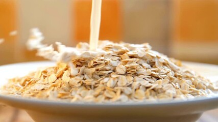 Slow motion closeup of milk pouring into a bowl of dry oatmeal. Preparing a healthy and nutritious breakfast cereal for a wholesome morning meal. - Powered by Adobe