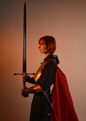 portrait of Fantasy warrior woman model, holding sword weapon, historical medieval armour costume with red fairytale cloak, strong fantasy knight. side profile pose on golden studio background.