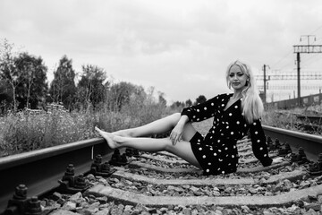 Young woman with long blonde hair, wearing a polka dot dress, sitting on railway tracks surrounded...