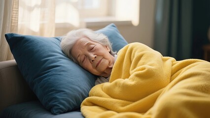 Elderly woman resting peacefully on a couch, covered with a yellow blanket and supported by a blue pillow.