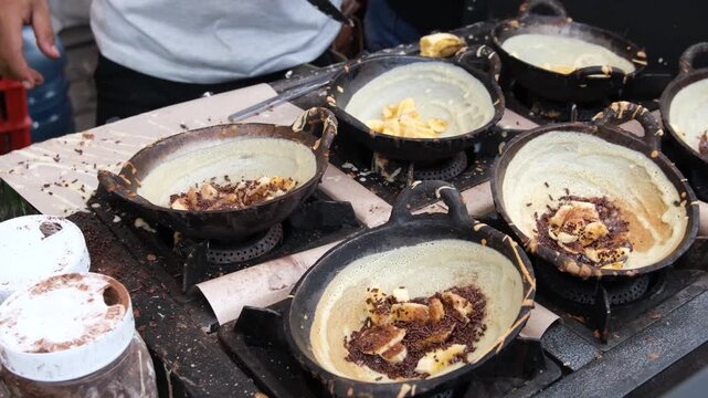 Street vendors cooking traditional Indonesian crepes called leker on multiple small pans.