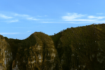 Rugged mountain landscape under a clear blue sky with wispy clouds. The steep slopes are covered in vegetation, contrasting with the exposed rock faces.